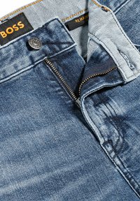 Vaqueros de mezclilla con un lavado azul, que presentan un zipper plateado, un botón negro y una etiqueta naranja. El material tiene una textura ligeramente rugosa con patrones de desgastado.