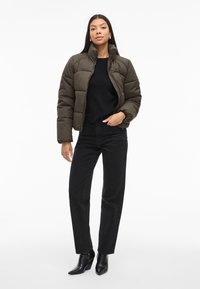 Giacca imbottita di colore verde oliva scuro, con colletto alto e texture trapuntata, indossata sopra un maglione nero e abbinata a jeans neri a vita alta.