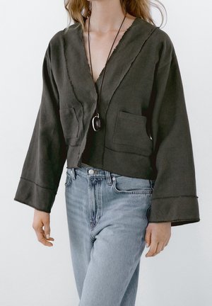 Femme portant une veste sombre ample avec deux poches avant, un collier pendentif et un jean taille haute bleu clair sur un fond uni.