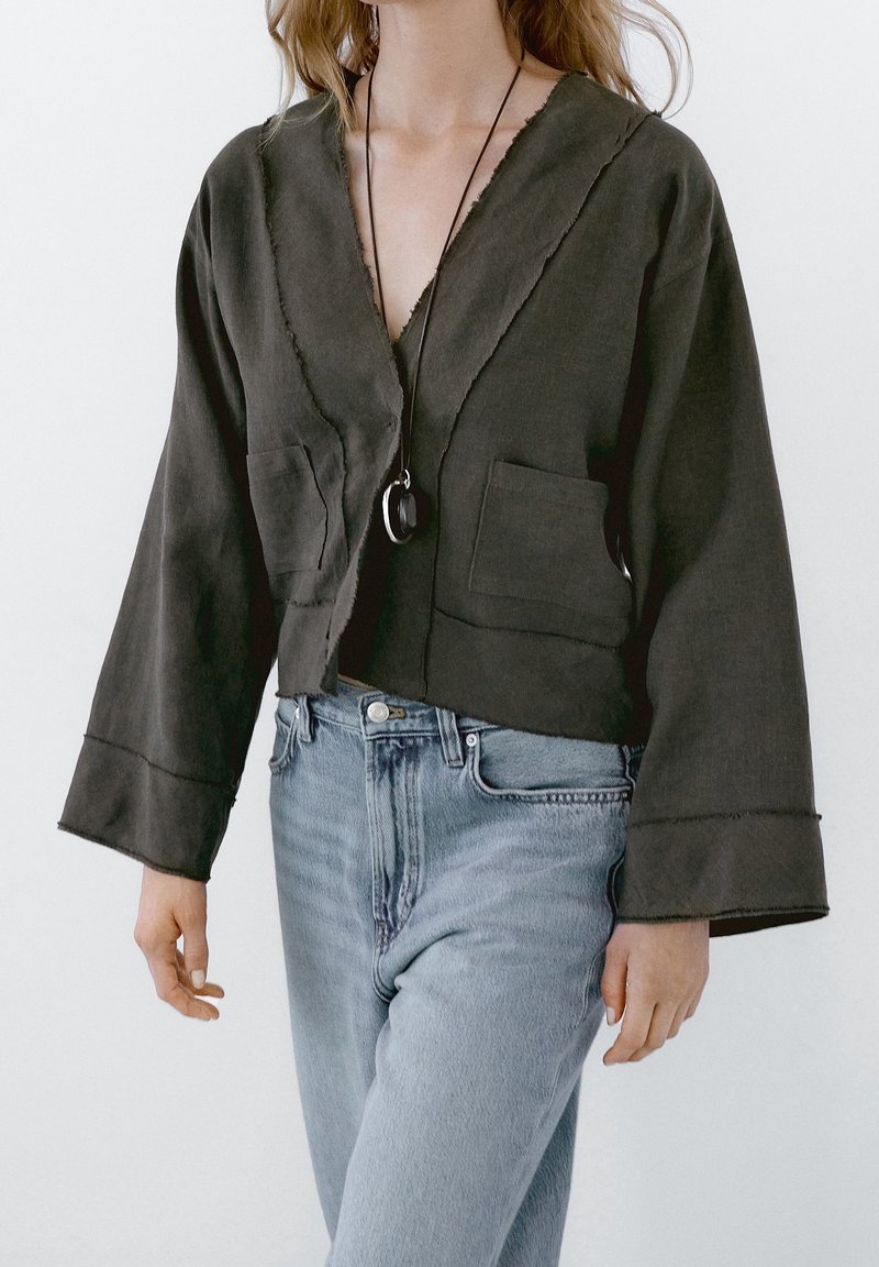 Femme portant une veste sombre ample avec deux poches avant, un collier pendentif et un jean taille haute bleu clair sur un fond uni.