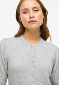 Lichtgrijze cardigan van zacht gebreid materiaal met een ronde halslijn, korte mouwen en vier knopen aan de voorkant.