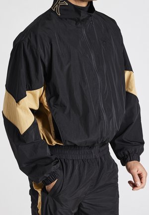 Homme portant une veste zippée noire et beige avec un pantalon assorti à taille élastique, mains partiellement dans les poches, debout devant un fond uni.