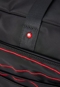Sac noir avec une surface texturée, des accents de couture rouge et un bouton-pression rouge proéminent sur une sangle. Compartiments zippés visibles.