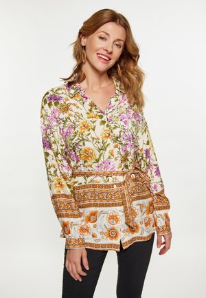 Frau lächelt, trägt eine Bluse mit floralem Muster mit orangefarbenen und violetten Blumen, kombiniert mit schwarzen Hosen vor einem schlichten weißen Hintergrund.