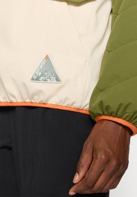 Chaqueta acolchada con secciones en color crema y verde oliva, ribete naranja y un parche triangular en el frente que muestra montañas y un sol.