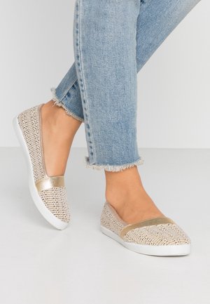 Chaussures slip-on tissées beige avec une bride métallique dorée et semelles blanches, portées avec un jean bleu clair à ourlet effiloché, jambes croisées.