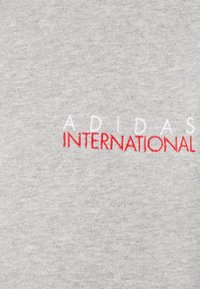 Grå bomull t-shirt med vit och röd broderad text "ADIDAS INTERNATIONAL" med en mjuk struktur och en rund halsdesign.