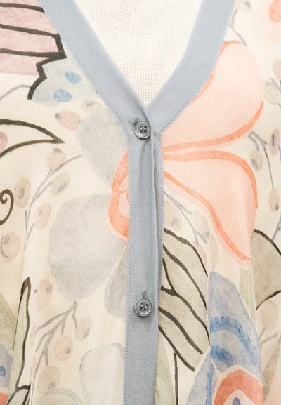 Κοντινή λήψη μπεζ ζακέτας με γαλάζιο περίγραμμα και κουμπιά, με μεγάλο αφηρημένο λουλουδάτο μοτίβο σε παστέλ ροζ, μπλε, γκρι και μαύρο.