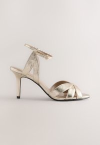 Metallisch goldene Sandalette mit hohem Absatz und Knöchelriemen, mit gekreuzten vorderen Bändern und einer glatten, glänzenden Textur.