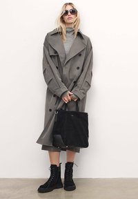 Trinchado gris de doble pecho con botones grandes, suéter ajustado de cuello alto, bolso tote de ante negro y botas negras con cordones y suelas gruesas.