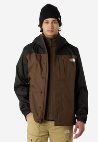 Brązowa i czarna wodoodporna kurtka z zamkiem z przodu i elastycznymi mankietami, w zestawieniu z brązową wewnętrzną warstwą na zamek i beżowymi spodniami.