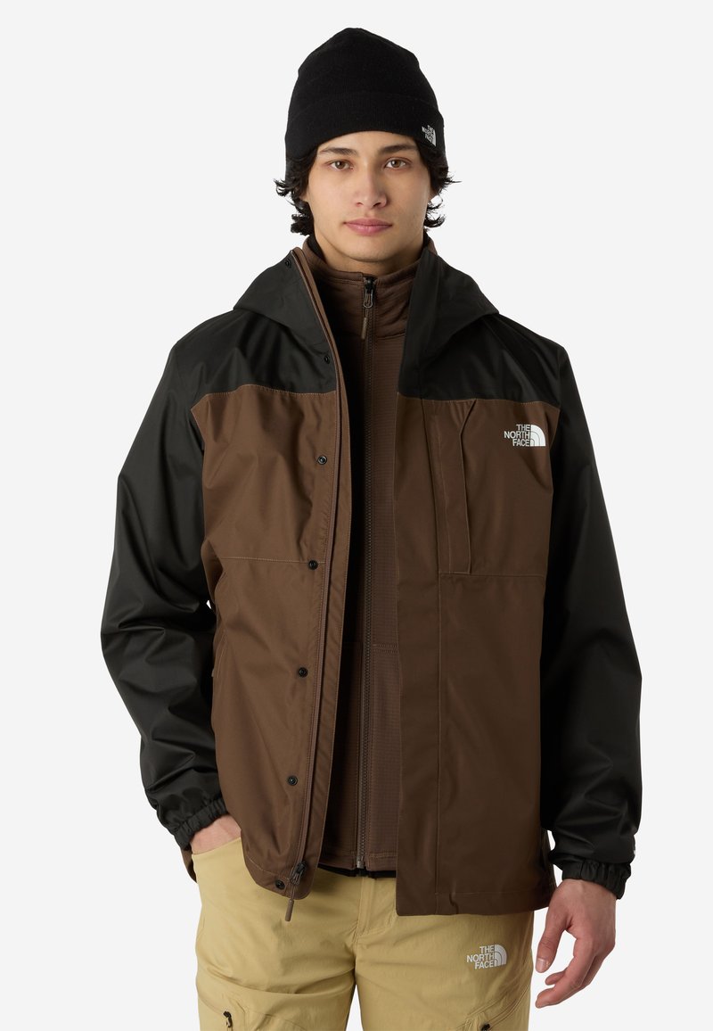 Brązowa i czarna wodoodporna kurtka z zamkiem z przodu i elastycznymi mankietami, w zestawieniu z brązową wewnętrzną warstwą na zamek i beżowymi spodniami.