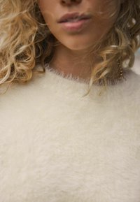 Fuzzy, cremefarvet sweater med en blød tekstur og ribbet halsudskæring. Detaljeret syning omkring kraven skaber en subtil kontrast.