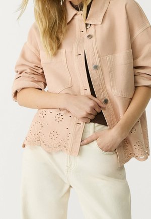 Femme portant une veste rose clair boutonnée avec un ourlet en dentelle ajourée, manches retroussées et pantalon crème taille haute, main dans la poche.