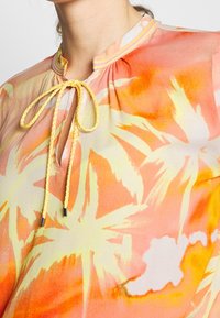 Kwiatowa bluzka w odcieniach pomarańczowym i żółtym, posiada dekolt w kształcie łezki z plecionym wiązaniem. Lekka tkanina o miękkiej, gładkiej fakturze.