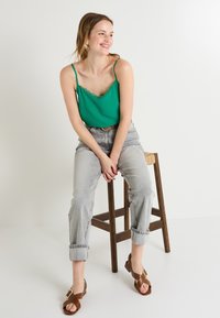 Camiseta verde con encaje, combinada con jeans de cintura alta en gris claro y sandalias morenas con tiras, sentada en un taburete de madera.