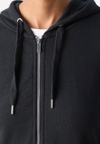 Gros plan d'une personne portant un sweat à capuche noir zippé sur une chemise blanche, montrant la fermeture éclair argentée et les cordons avec embouts métalliques.