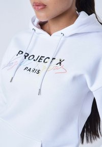 Witte hoodie met een vooraanstaand graphic van "PROJECT X PARIS" in verschillende vetgedrukte lettertypes. Koorden met metalen tips en een zachte, textuurstof.