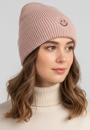 Pink ribbed knit beanie with a smiley face patch. Soft texture, slouchy design, and features a folded brim. Worn with a beige turtleneck.