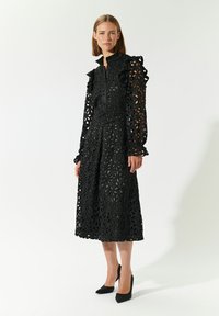 Schwarzes Kleid mit detailliertem durchbrochenem Blumenmuster, langen Rüschensäumen, hohem Kragen und taillierter Taille. Kombiniert mit schwarzen hohen Schuhen.