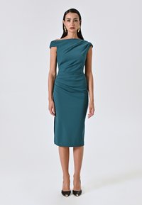 Türkises, figurbetontes Kleid mit schulterfreiem Design, Raffung, knielanger Länge, seitlichem Schlitz und glattem Stoff. Schwarze spitze Absätze.