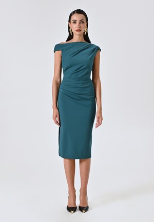 Türkises, figurbetontes Kleid mit schulterfreiem Design, Raffung, knielanger Länge, seitlichem Schlitz und glattem Stoff. Schwarze spitze Absätze.