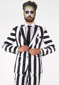 Čierno-biely pruhovaný oblek s lesklým povrchom, obsahujúci sako a zodpovedajúce nohavice; model má živú zeleno-bielu tvárovú maľbu.