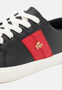 Mustat nahkakengät punaisella raidalla, joissa on kultainen logo, valkoiset nauhat ja teksturoidut kumipohjat.