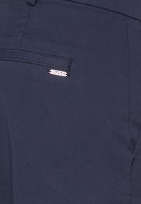 Tmavě modré kalhoty s hladkým povrchem, s malým stříbrným logem "HUGO" na zadní kapse. Klasický design s rovnými nohavicemi.