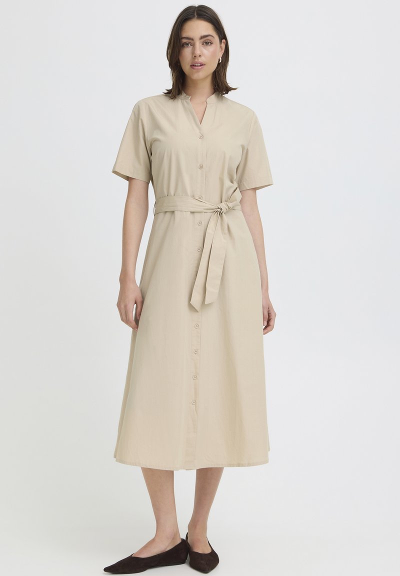 Femme portant une robe midi beige à manches courtes avec des boutons sur le devant, une ceinture nouée à la taille et des chaussures plates noires, debout devant un fond uni.