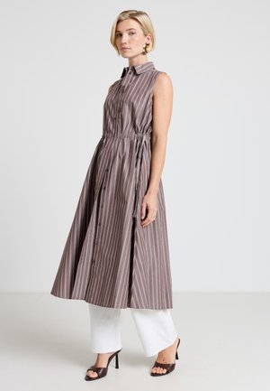 Donna con capelli corti e biondi che indossa un vestito midi marrone a righe senza maniche sopra pantaloni bianchi a gamba larga e sandali neri con il tacco, in piedi.