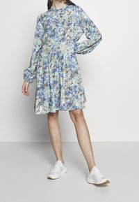 Robe fleurie aux teintes bleues, vertes et marron, dotée de manches longues, d'un col rond et d'une jupe à volants. Associée à des baskets blanches.