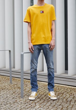 Persoon die een gele Tommy Jeans T-shirt, een blauwe spijkerbroek en gele sneakers draagt, staand op een kasseienstraat vlakbij metalen fietsenrekken.