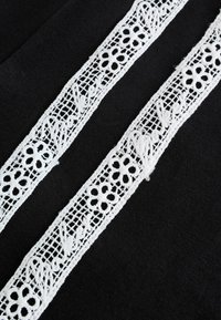 Două panglici albe din dantelă, paralele, cu modele florale și de frunze pe un fundal de material negru.