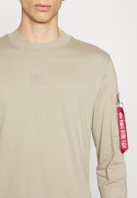 Vaaleanbeige pitkähihainen paita, jossa on pyöreä kaula-aukko. Paidassa on teksturoitu logopatches rintataskussa ja sivussa vetoketjutasku punaisella vetoketjulla.