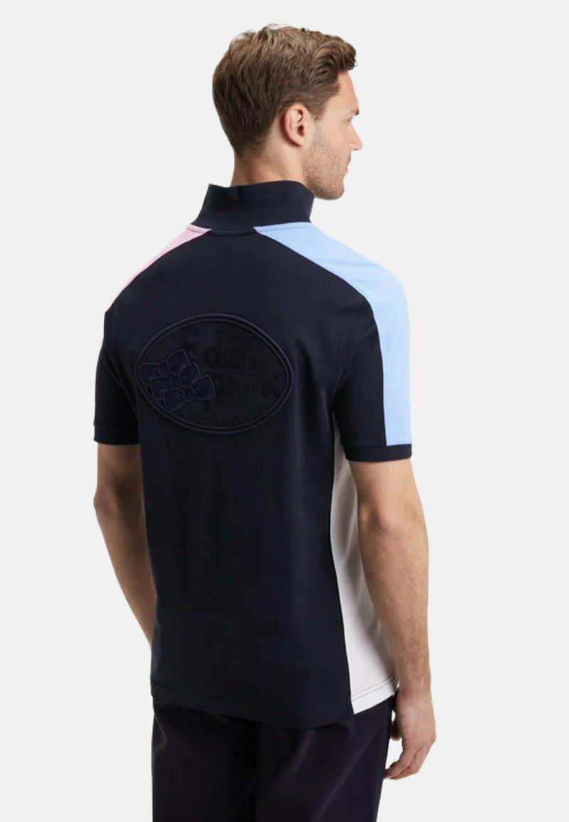 Homme portant un polo bleu marine à manches courtes et col haut avec des blocs de couleur rose, bleu et blanc, tourné de dos sur un fond uni.