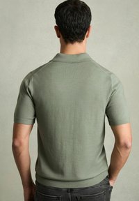 Tricou polo cu mânecă scurtă, de culoare verde măslinie, realizat dintr-un material neted, cu guler și tiv cu țesătură în relief. Are un croi ajustat.
