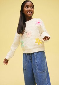 Pulover alb tricotat cu broderie colorată în formă de flori; asociat cu blugi din denim blua în talie înaltă. Fundal galben deschis.