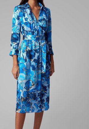 Mujer con un vestido midi azul con estampado floral, con cuello, mangas tres cuartos y un cinturón de tela a juego atado a la cintura.
