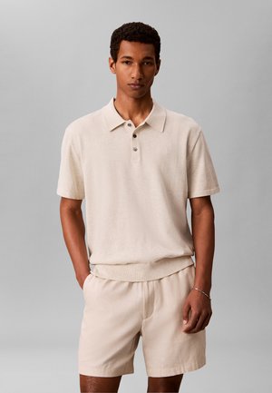 Mężczyzna ubrany w beżową koszulkę polo z krótkim rękawem i pasujące szorty, stojący z jedną ręką w kieszeni na jednolitym szarym tle.
