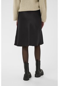 Jupe en satin noir avec une silhouette fluide, associée à des bottes de combat noires texturées et des collants noirs transparents. Pull oversize beige clair.