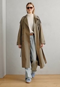 Kaki trenchcoat med dobbeltradede knapper, kombinert med en hvit ribbestrikket genser og lys blå jeans, iført grå sneakers og solbriller.