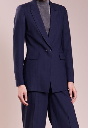 Blazer bleu marine à double boutonnage avec deux poches avant, revers aigus et coupe ajustée. Porté par-dessus un col roulé gris texturé.