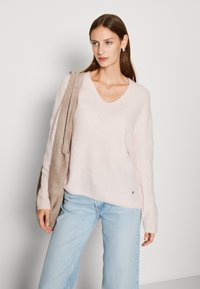 Lichtroze v-hals trui van zacht materiaal met een losse pasvorm en ribbeltextuur, gecombineerd met lichte spijkerbroek en een beige schoudertas.