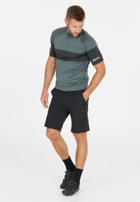 Herren-Sportoutfit mit einem graugrünen Kurzarmoberteil mit schwarzen Akzenten und schwarzen Shorts. Inklusive seitlicher Reißverschlusstasche und Radschuhen.