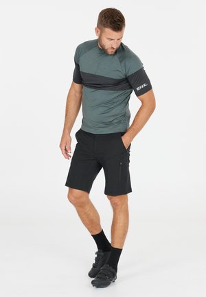 Herren-Sportoutfit mit einem graugrünen Kurzarmoberteil mit schwarzen Akzenten und schwarzen Shorts. Inklusive seitlicher Reißverschlusstasche und Radschuhen.