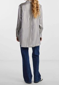 Blusa lunga manica in satin argentato con piega sul retro, abbinata a jeans larghi blu scuro e un accenno di colletto.