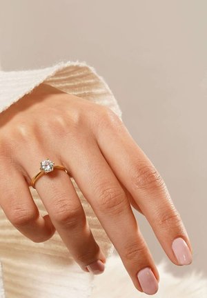 Gold ring with a round, clear gemstone centerpiece, set on a smooth band. Hand appears relaxed, with light pink nail polish.
