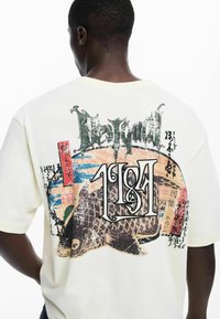 Camiseta de color crema con un estampado gráfico de un pez, texto superpuesto y acentos coloridos, con mangas cortas y un ajuste relajado.
