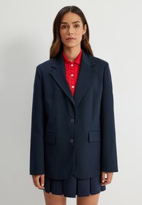 Granatowa marynarka z strukturalnego materiału, z klapami w szpic, na dwa guziki i kieszeniami z przodu; nałożona na czerwony sweter w prążki.